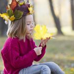 A Girl Examines A Maple Leaf. Child On A Walk In A Crown Of Leaves. Preschool Girl In The Autumn Park.