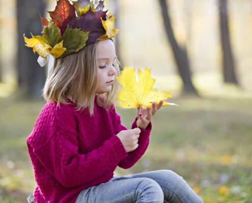 A Girl Examines A Maple Leaf. Child On A Walk In A Crown Of Leaves. Preschool Girl In The Autumn Park.