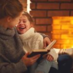 Happy Family Mother And Child Daughter Read Book On Winter Evening Near Fireplace