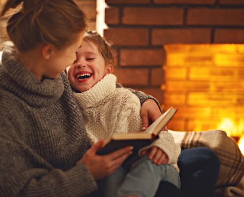 Happy Family Mother And Child Daughter Read Book On Winter Evening Near Fireplace